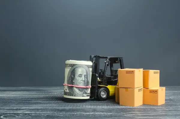 A miniature forklift carrying a roll of money next to cardboard boxes, illustrating the cost factors for shipping from Dubai to Bahrain.